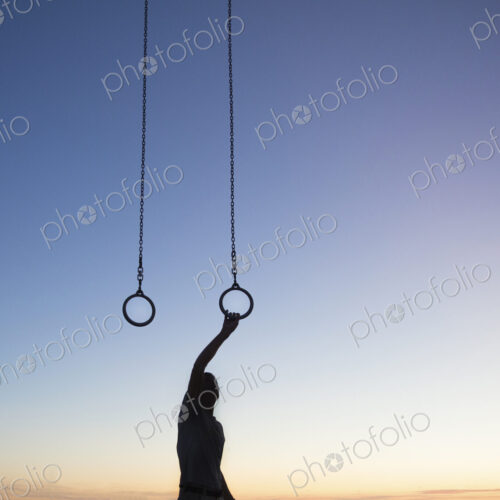 Silhouette of man standing outdoors at sunset, arm raised, holding onto gymnastic rings. Silhouette of man standing outdoors at sunset, arm raised, holding onto gymnastic rings.