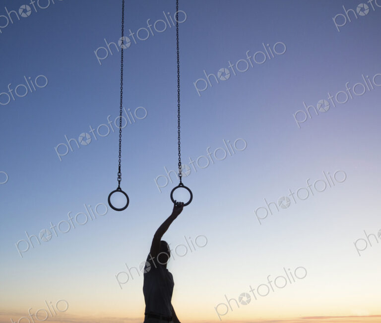 Silhouette of man standing outdoors at sunset, arm raised, holding onto gymnastic rings.