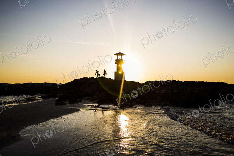 Silhouette of two people walking past lighthouse by the ocean at sunset.