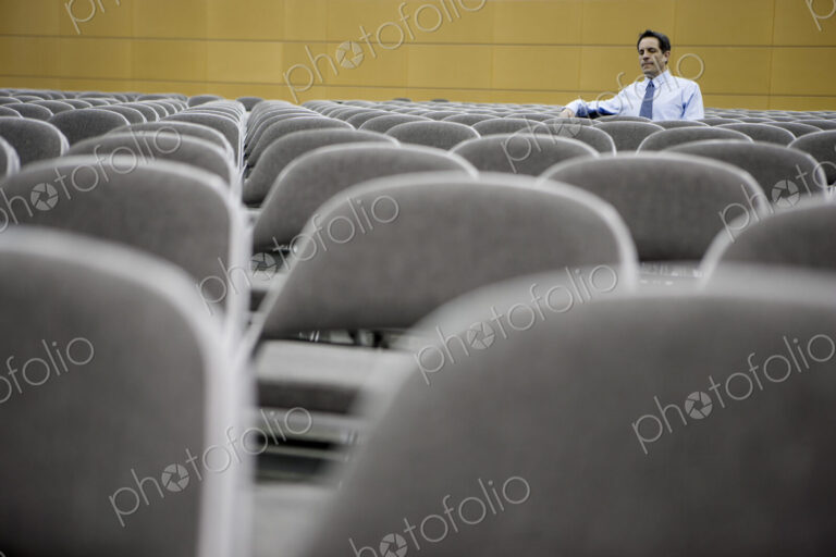 Single businessman sitting in a room full of chairs prior to a conference centre meeting.