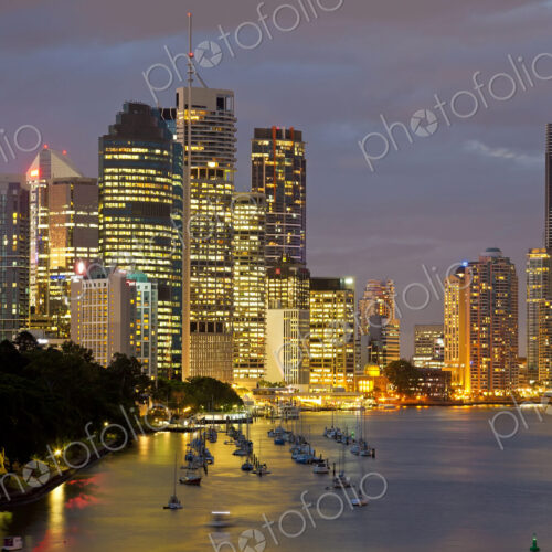 Skyline of city by the ocean at dusk, with illuminated skyscrapers, sailing boats…