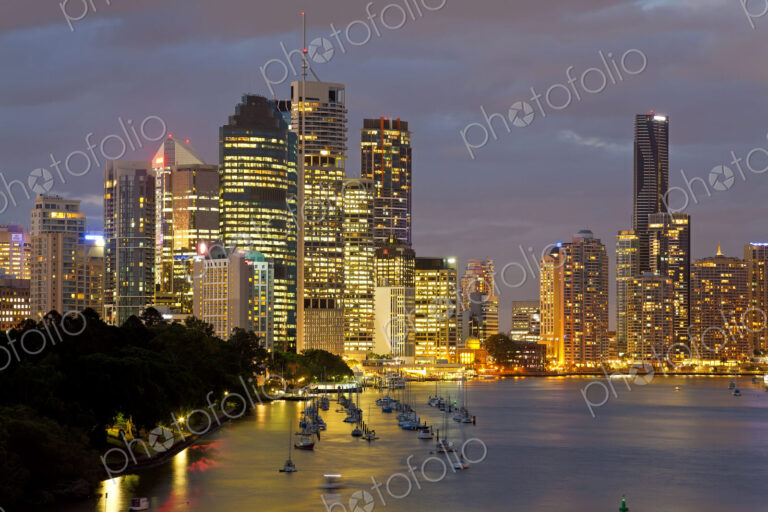 Skyline of city by the ocean at dusk, with illuminated skyscrapers, sailing boats…