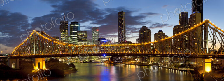 Skyline of city, with illuminated skyscrapers and bridge over river in the foreground.