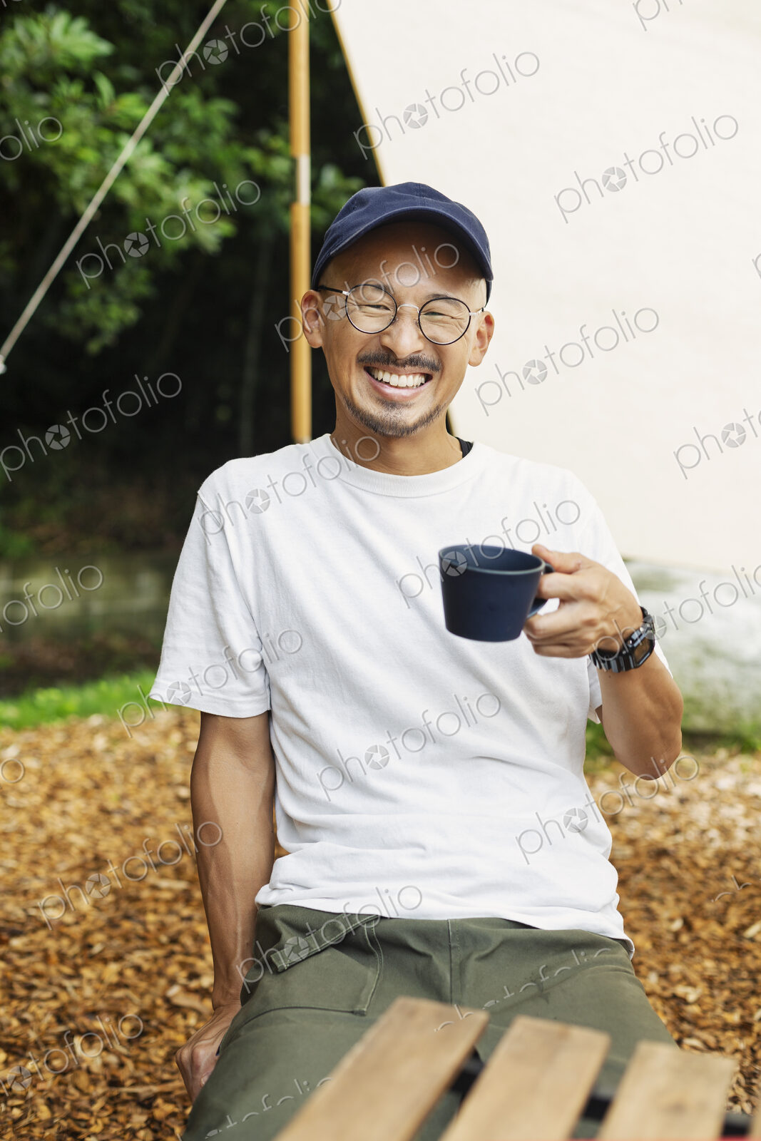 Smiling Japanese man wearing baseball cap and glasses sitting outdoors at a table,…