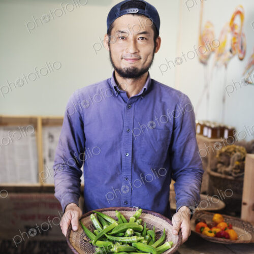 Smiling Japanese man wearing cap standing in farm shop, holding bowl with fresh okra. Smiling Japanese man wearing cap standing in farm shop, holding bowl with fresh okra.