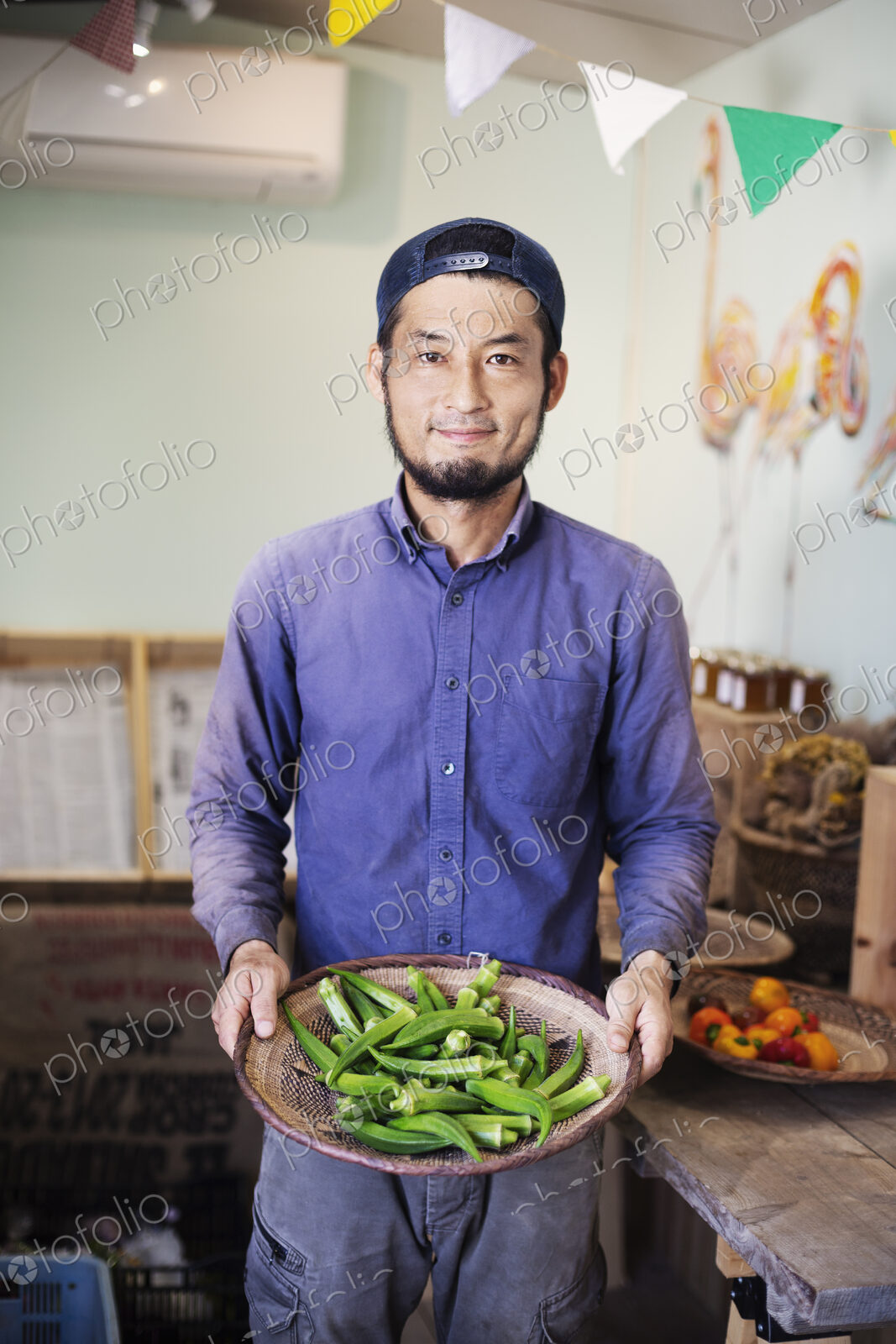 Smiling Japanese man wearing cap standing in farm shop, holding bowl with fresh okra.