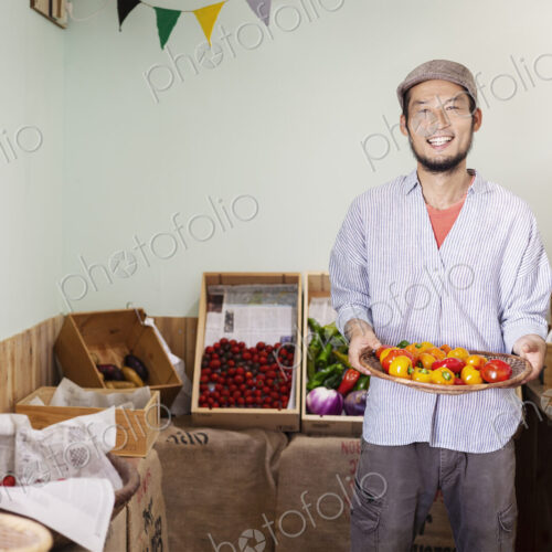Smiling Japanese man wearing cap standing in farm shop, holding bowl with fresh…