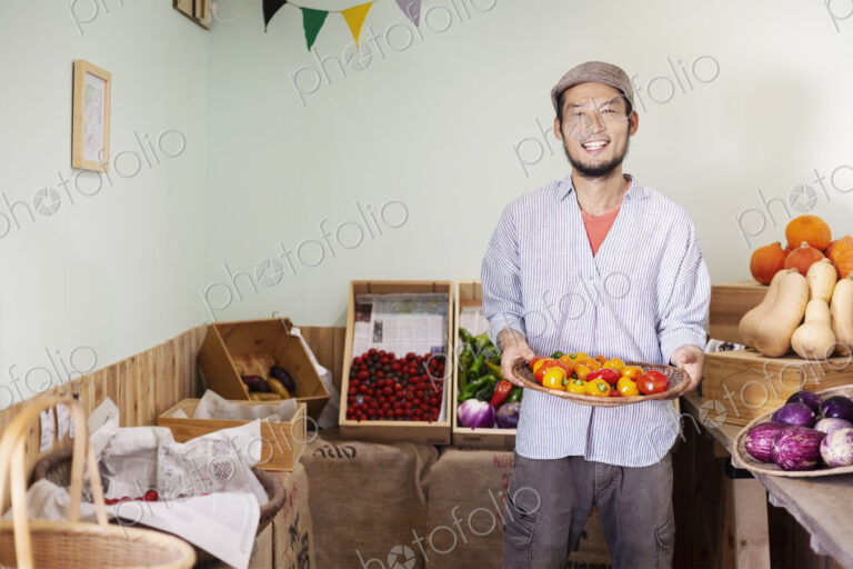 Smiling Japanese man wearing cap standing in farm shop, holding bowl with fresh…