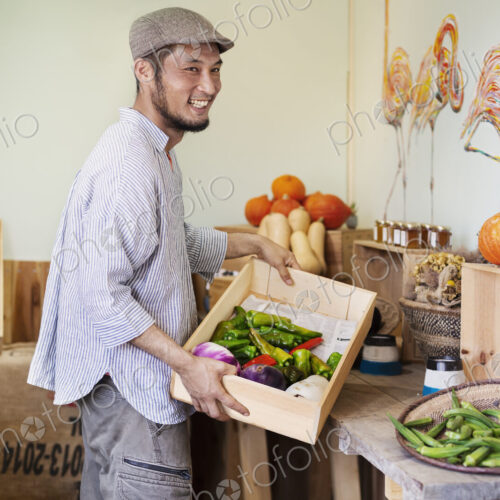 Smiling Japanese man wearing cap standing in farm shop, holding crate with fresh… Smiling Japanese man wearing cap standing in farm shop, holding crate with fresh…