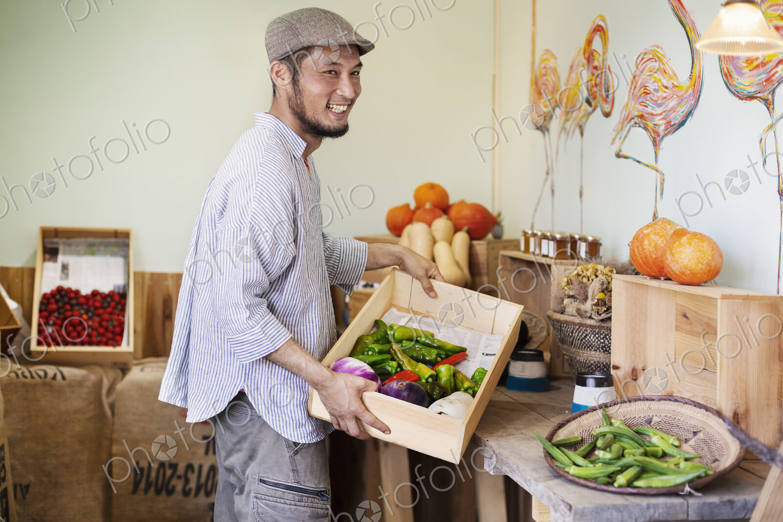 Smiling Japanese man wearing cap standing in farm shop, holding crate with fresh…