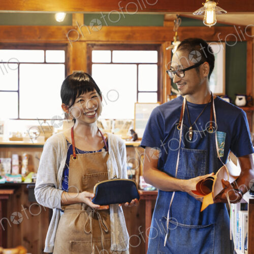 Smiling Japanese woman and man wearing blue apron and glasses standing in a leather shop. Smiling Japanese woman and man wearing blue apron and glasses standing in a leather shop.