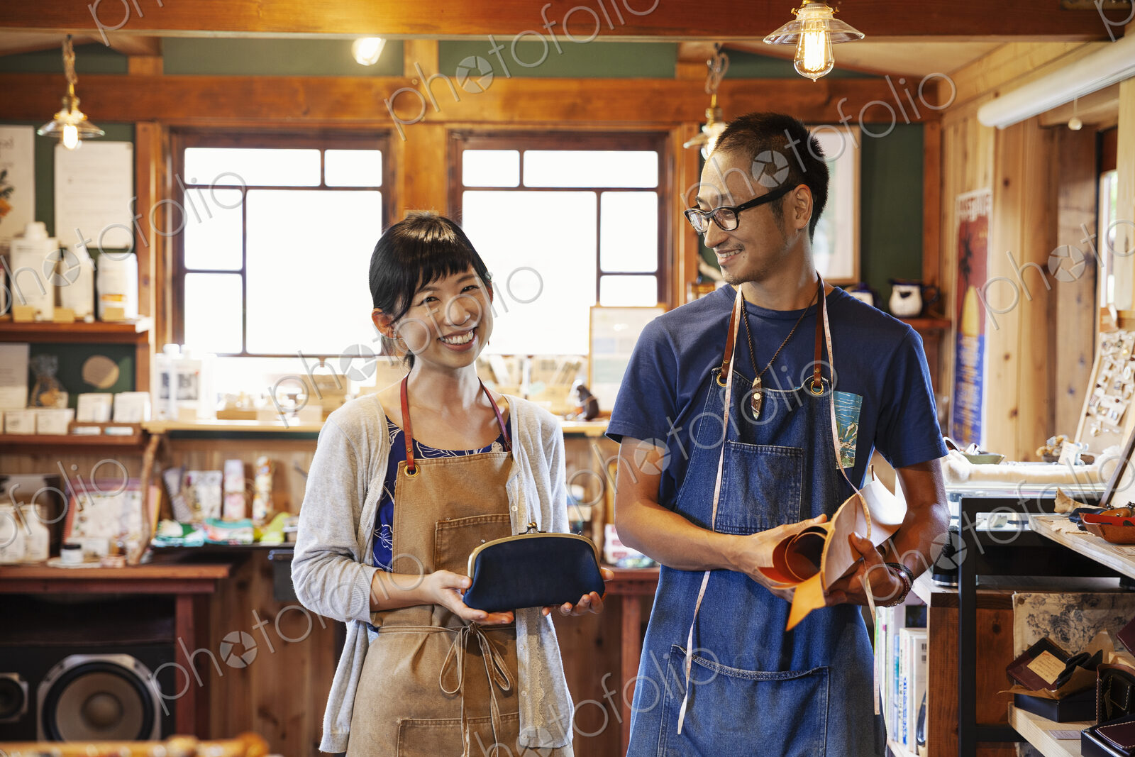 Smiling Japanese woman and man wearing blue apron and glasses standing in a leather shop.