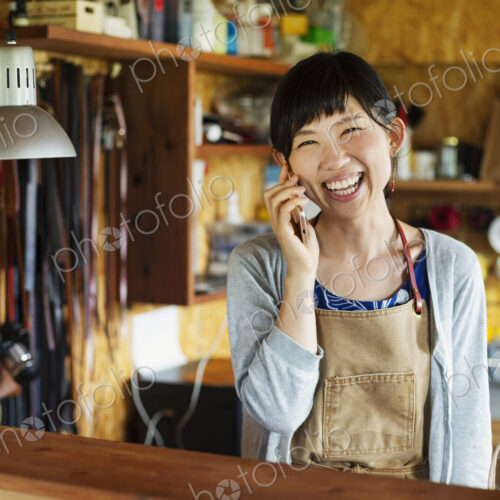 Smiling Japanese woman wearing apron standing in a leather shop, talking on her mobile…