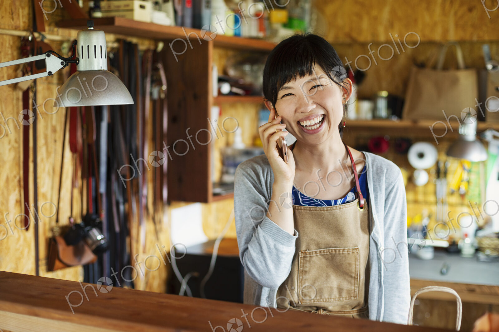 Smiling Japanese woman wearing apron standing in a leather shop, talking on her mobile…