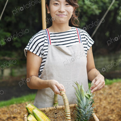 Smiling Japanese woman wearing apron standing outdoors, holding basket with fresh… Smiling Japanese woman wearing apron standing outdoors, holding basket with fresh…