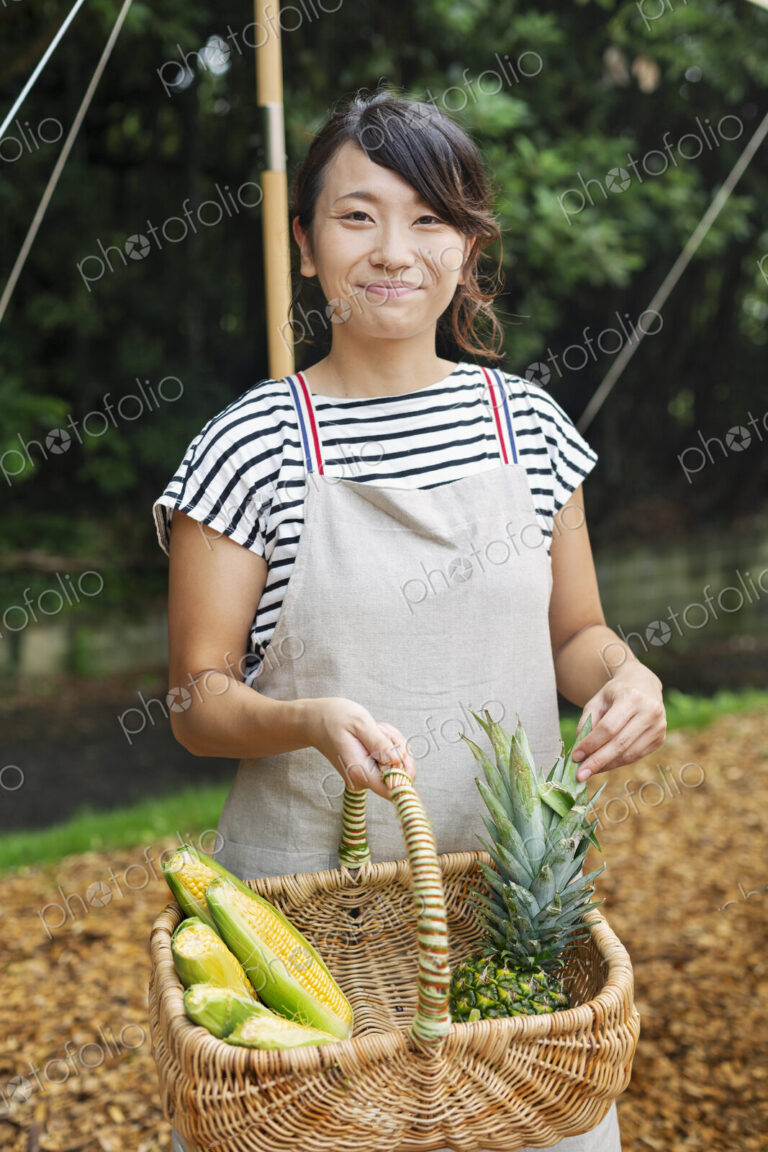 Smiling Japanese woman wearing apron standing outdoors, holding basket with fresh…