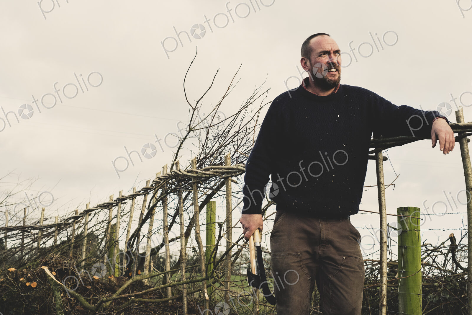 Smiling bearded man holding axe and bill hook standing next to a newly built…