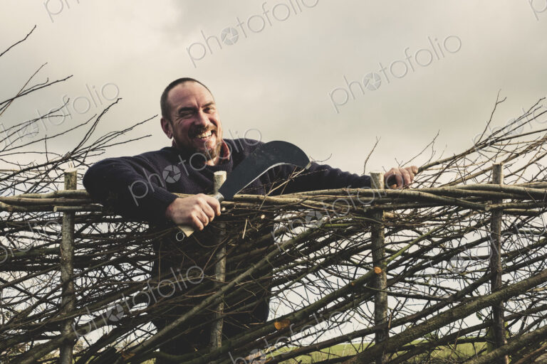 Smiling bearded man holding bill hook standing next to a newly built traditional hedge.