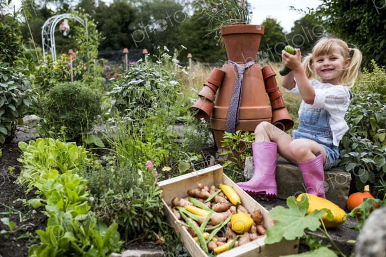 Smiling blond girl sitting in garden next to terracotta scarecrow, picking fresh…