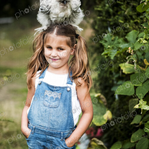 Smiling blond girl standing in a garden, with a fluffy grey chicken on her head. Smiling blond girl standing in a garden, with a fluffy grey chicken on her head.