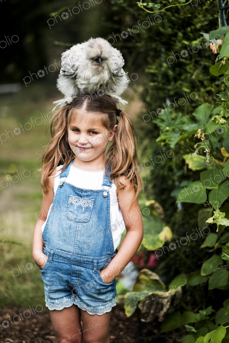 Smiling blond girl standing in a garden, with a fluffy grey chicken on her head.