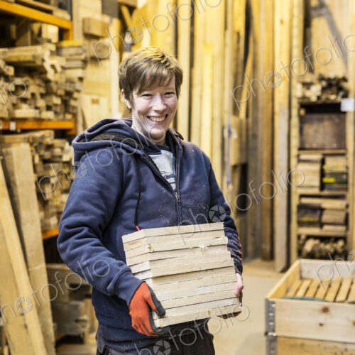 Smiling blond woman wearing work gloves standing in a workshop, holding stack of wood,… Smiling blond woman wearing work gloves standing in a workshop, holding stack of wood,…