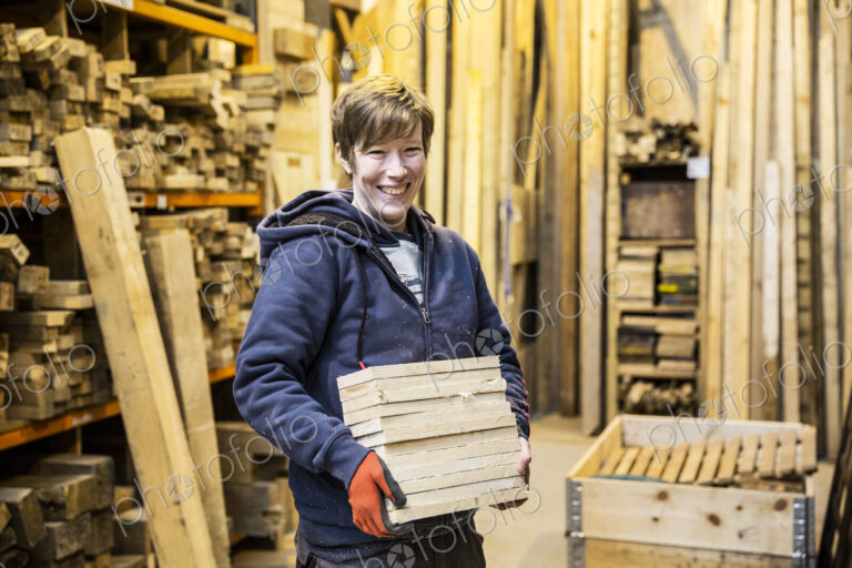 Smiling blond woman wearing work gloves standing in a workshop, holding stack of wood,…