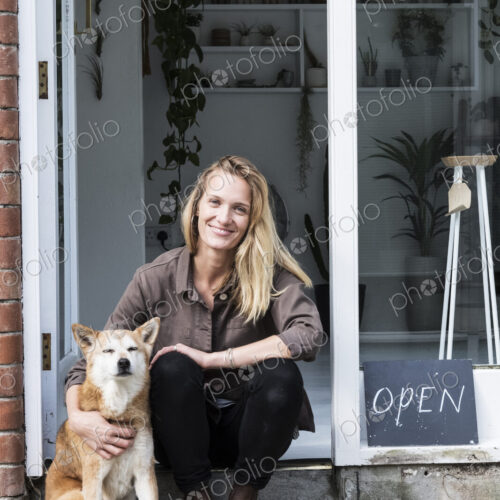 Smiling female owner of plant shop sitting on steps outside her store, a dog sitting…