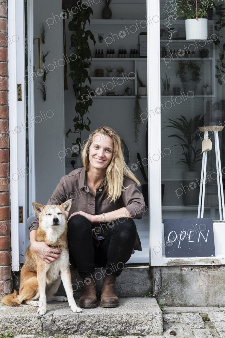 Smiling female owner of plant shop sitting on steps outside her store, a dog sitting…