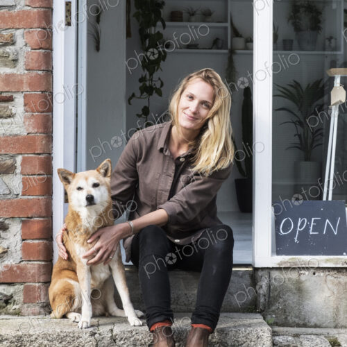 Smiling female owner of plant shop sitting on steps outside her store, a dog sitting…
