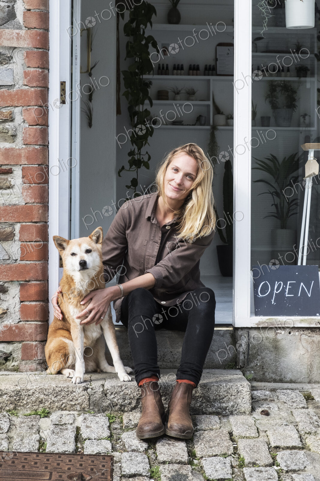 Smiling female owner of plant shop sitting on steps outside her store, a dog sitting…