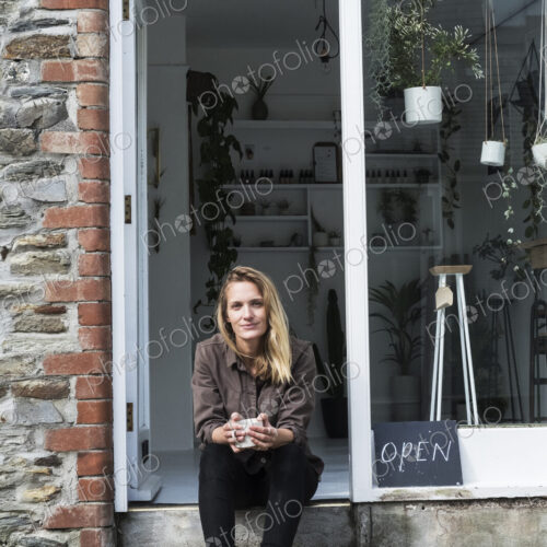 Smiling female owner of plant shop sitting on steps outside her store, looking at camera.