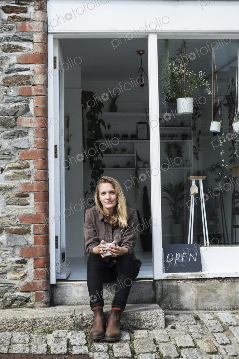 Smiling female owner of plant shop sitting on steps outside her store, looking at camera.