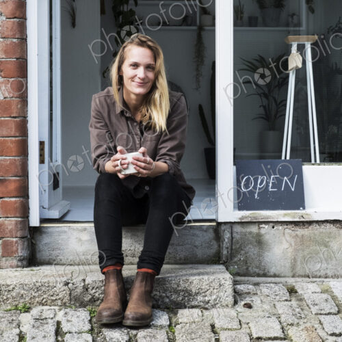 Smiling female owner of plant shop sitting on steps outside her store, looking at camera.