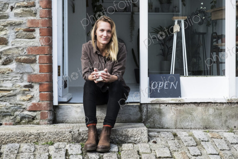 Smiling female owner of plant shop sitting on steps outside her store, looking at camera.