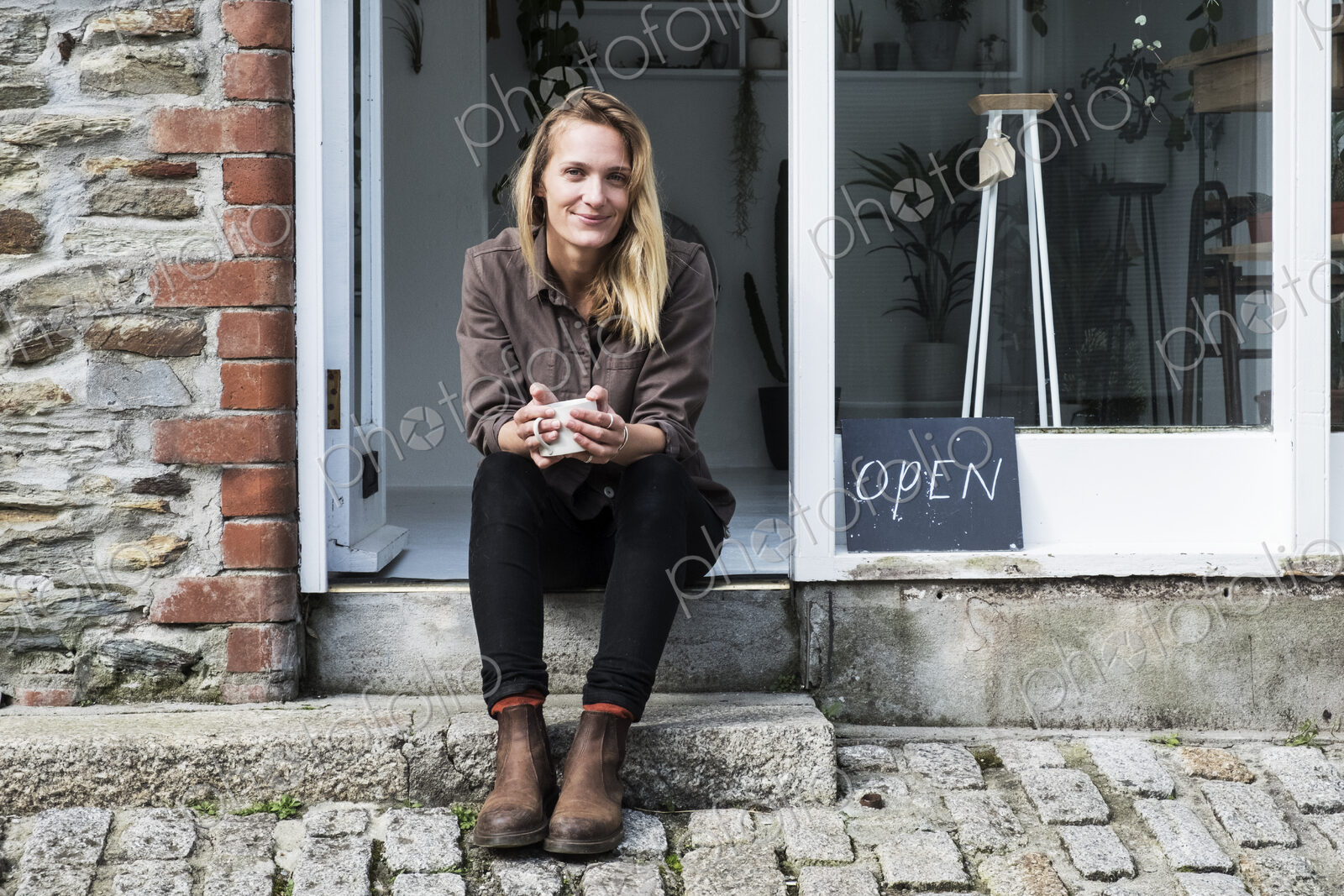 Smiling female owner of plant shop sitting on steps outside her store, looking at camera.