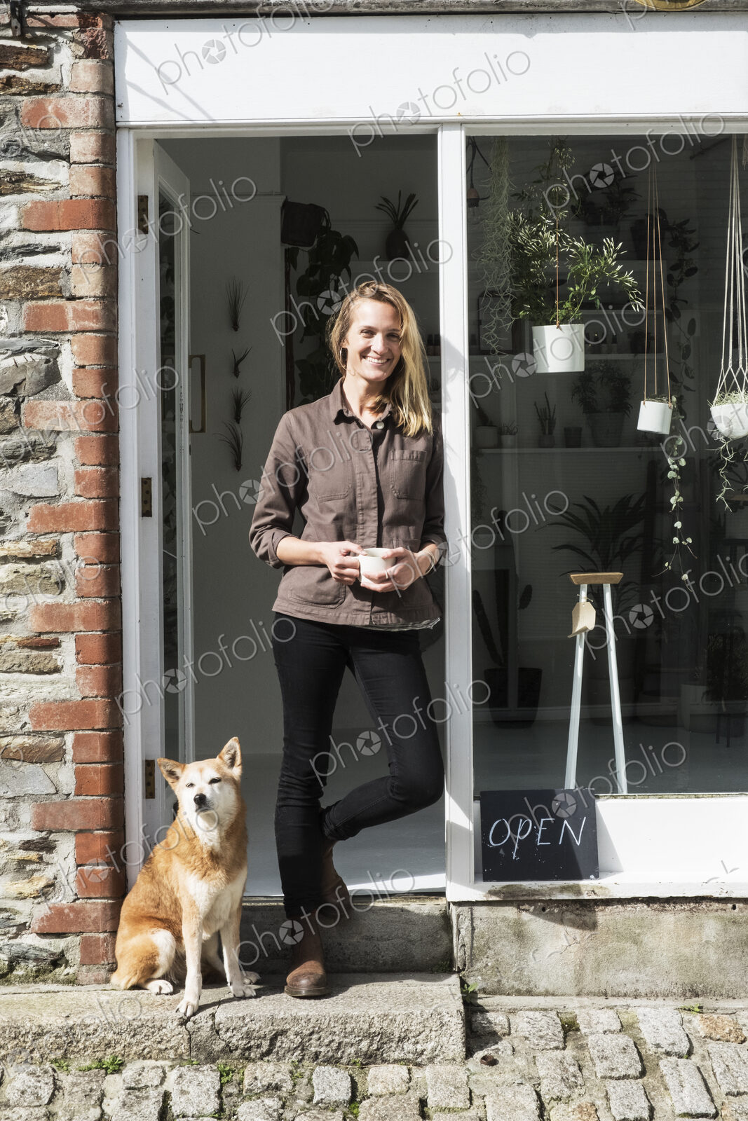Smiling female owner of plant shop standing outside her store, a dog sitting next to her.