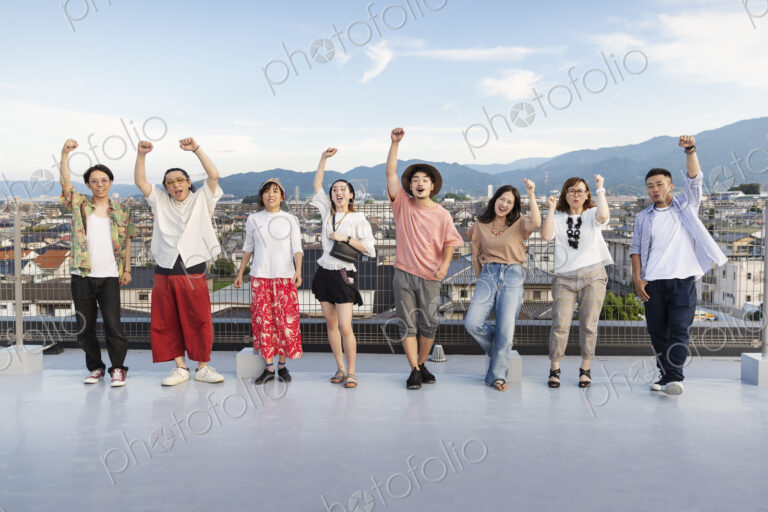 Smiling group of young Japanese men and women standing on a rooftop in an urban setting.