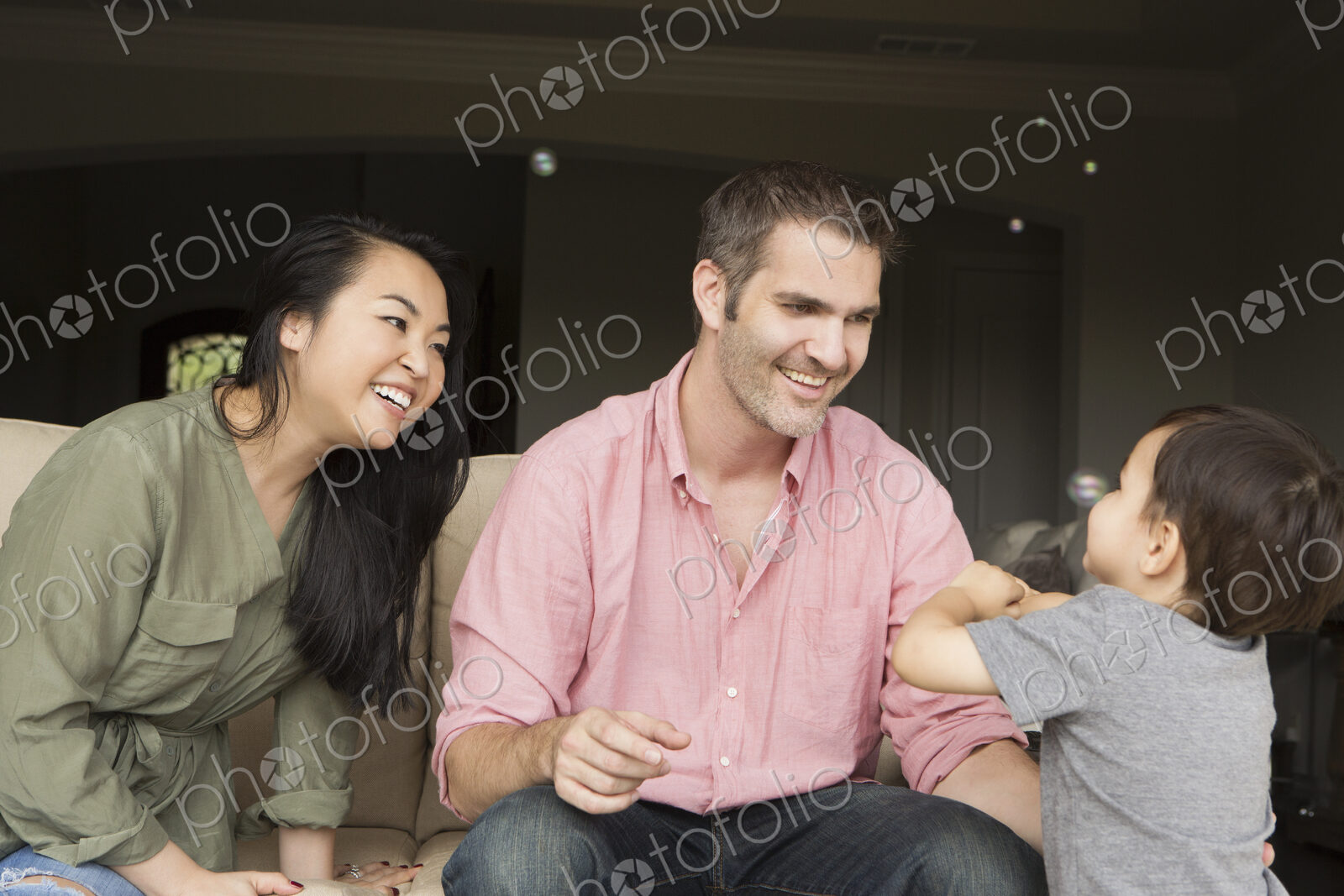 Smiling man and woman sitting side by side on a sofa, playing with their young son.
