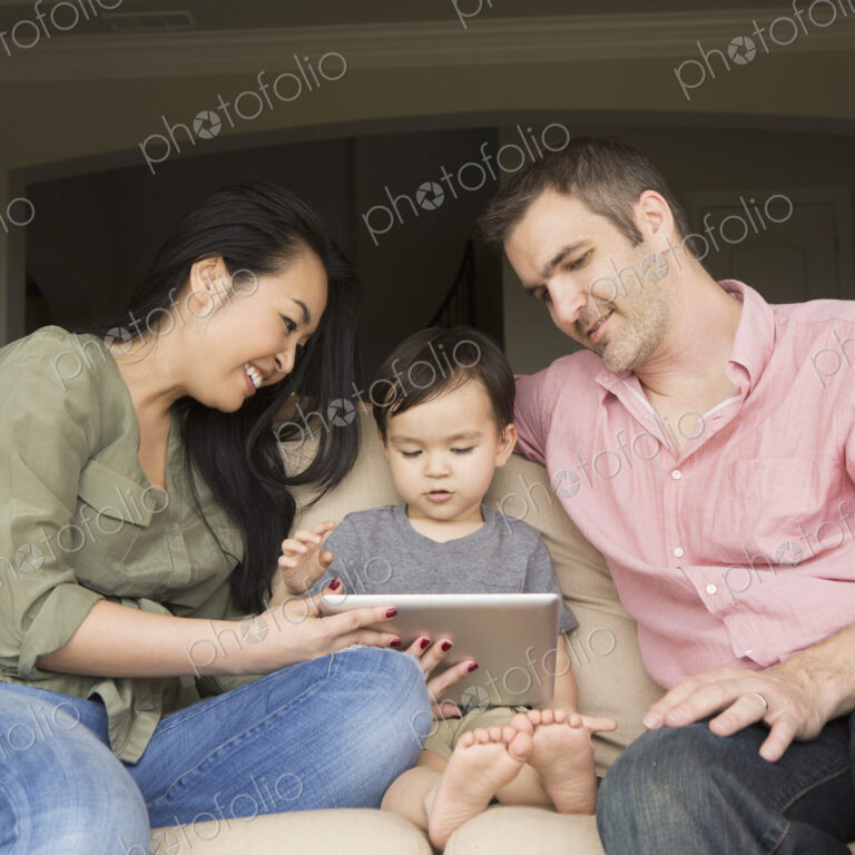 Smiling man and woman sitting side by side on a sofa with their young son, looking at…