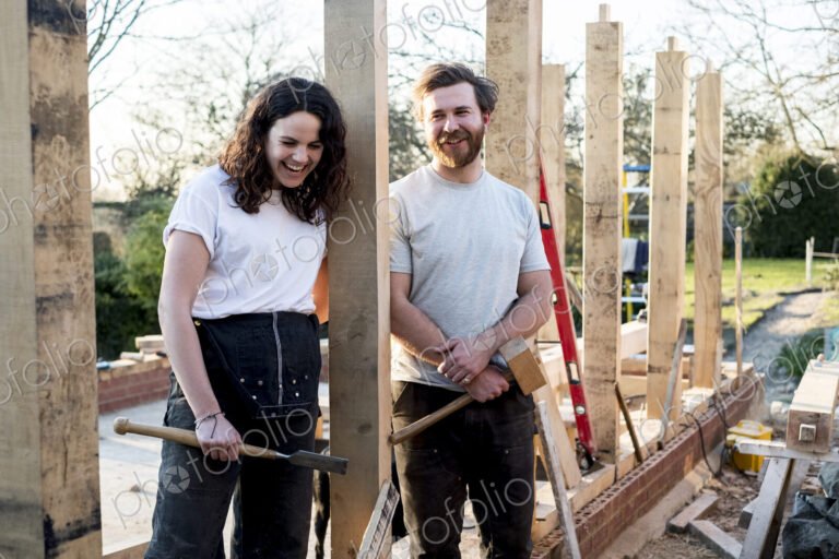 Smiling man and women holding hand tools standing on building site of residential…