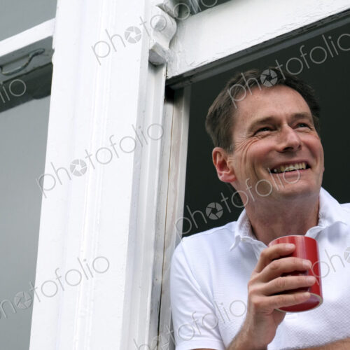 Smiling man leaning out of window holding hot drink.