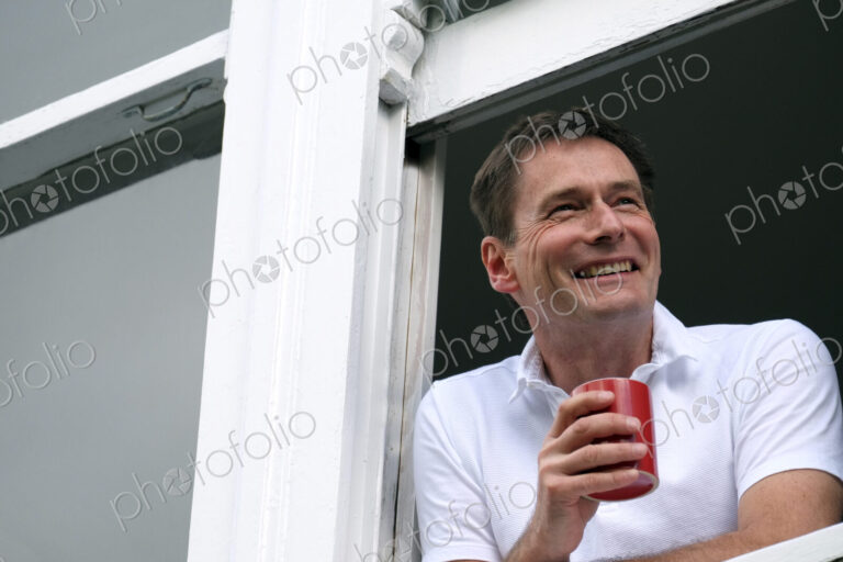 Smiling man leaning out of window holding hot drink.