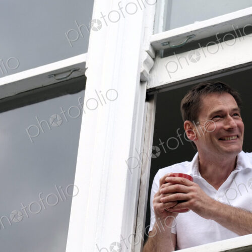 Smiling man leaning out of window holding hot drink.