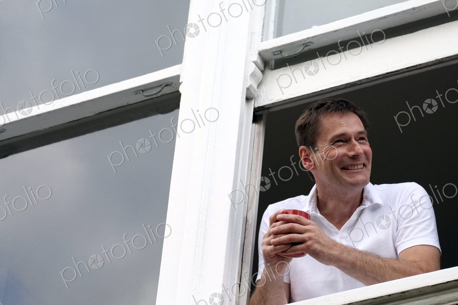 Smiling man leaning out of window holding hot drink.