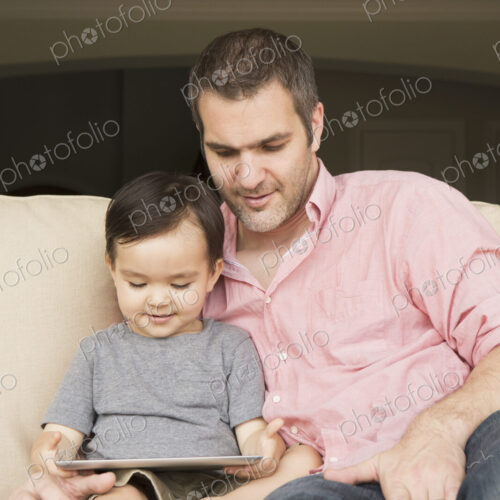 Smiling man sitting on a sofa with his young son, looking at a digital tablet. Smiling man sitting on a sofa with his young son, looking at a digital tablet.