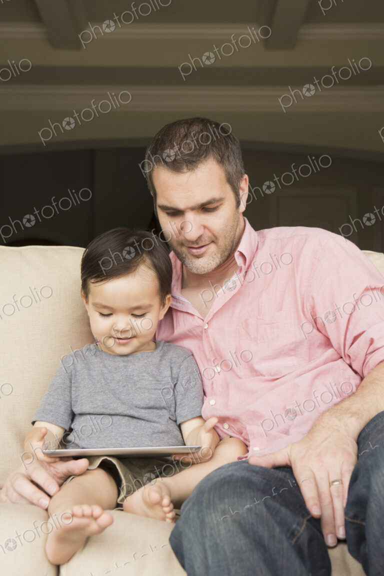 Smiling man sitting on a sofa with his young son, looking at a digital tablet.