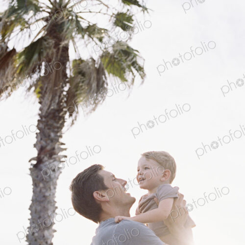 Smiling man standing by a palm tree, carrying his young son in his arms.