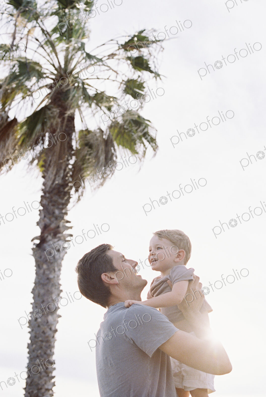 Smiling man standing by a palm tree, carrying his young son in his arms.