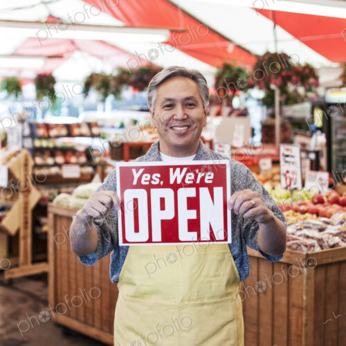 Smiling man wearing apron at a fruit and vegetable market, holding aloft Open sign..